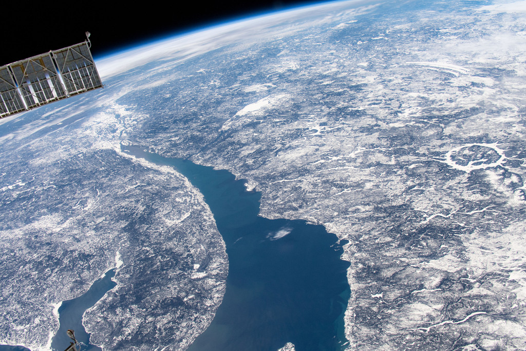 Der Blick von der Internationalen Raumstatzion ISS fällt auf die Erde. In der Mitte verläuft ein breites Gewässer, rechts auf halber Bildhöhe ist eine sehr markante weiße runde Struktur, die vermutlich von einem Meteoriteneinschlag stammt. Es ist der Manicouagankrater.