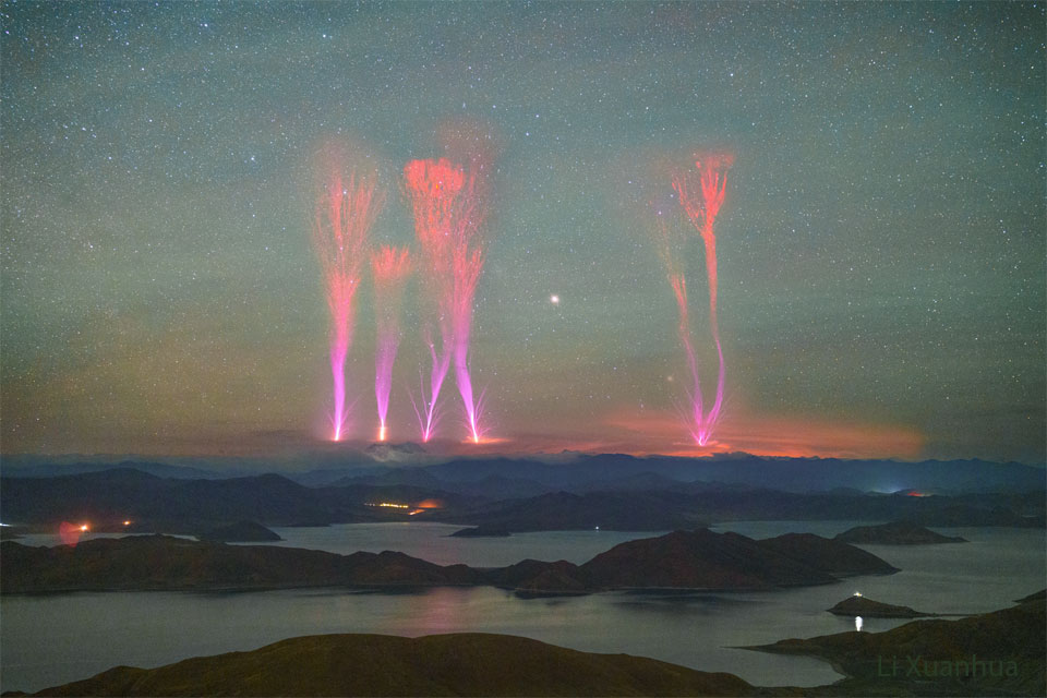 Hinter einer Landschaft mit Bergen und Wasser leuchten vier gewaltige rote Strahlenblitze, sogenannte Red Sprites oder Rote Kobolde. Unten sind dichte helle Blitze, die sich nach oben hin in rote, stark verästelte Büschel auffächern.