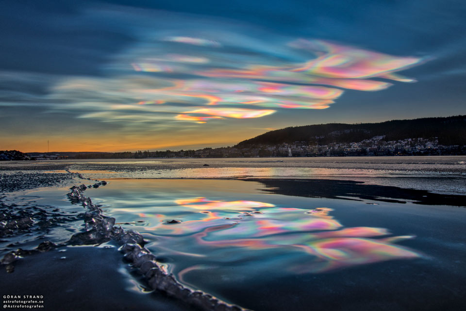 Bunt schillernde Wolken spiegeln sich im Wasser an einem Ufer, am hinteren Gewässerrand ist eine Stadt und eine bewaldete Anhöhe.