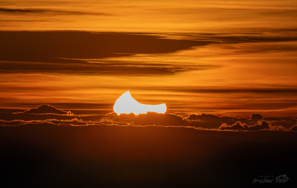Am orange beleuchteten Himmel steigt die Sonne hinter den Wolken auf. Sie ist teilweise vom Mond verdeckt und hat daher eine Sichelform.