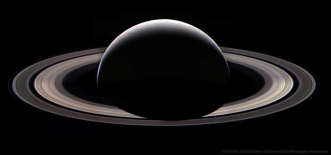 Der Planet Saturn ist von der Nachtseite aus schräg von oben zu sehen. Die Ringe sind beleuchtet, vom Planeten sieht man eine nach oben gerichtete Sichel, er wirft einen Schatten auf die Ringe.
