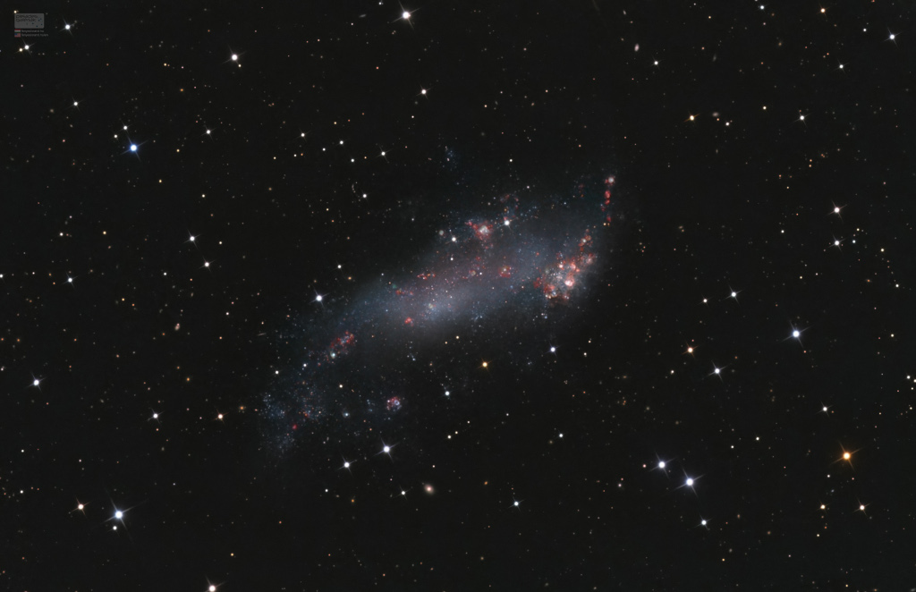 Mitten im Bild schwebt die kleine Galaxie IC 2574 hinter lose verteilten Sternen. Sie hat viele rötliche Sternbildungsregionen, die man an ihrem roten Leuchten erkennt.