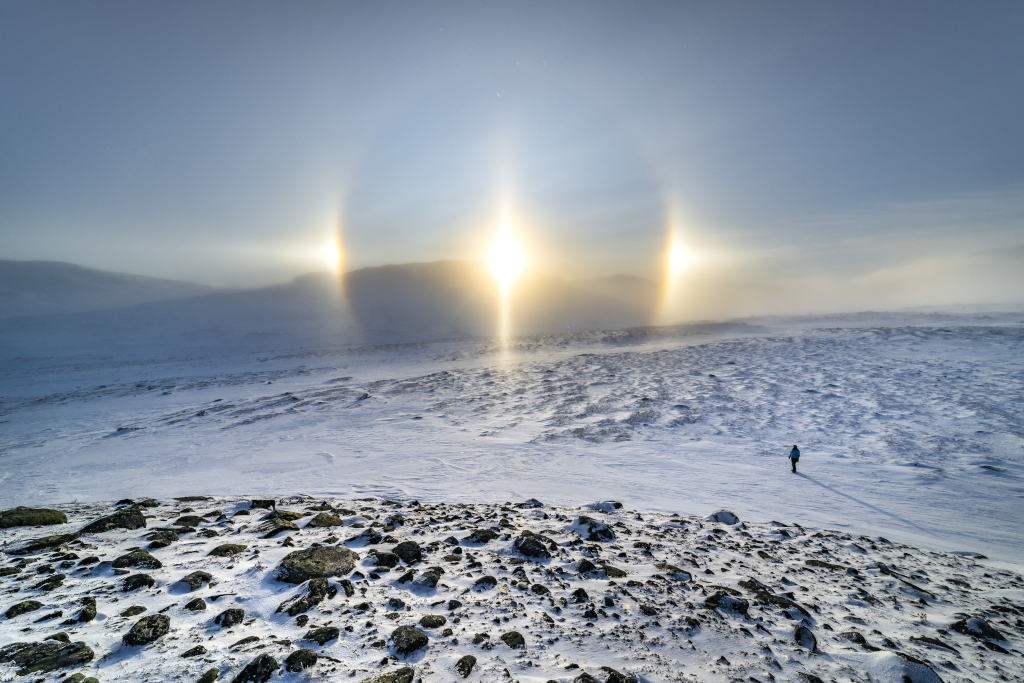 Über einem verschneiten Feld mit einer einsamen Person leuchtet über einem Hügel am Horizont die Sonne. Sie ist von einem runden Hof umgeben, links und rechts leuchten helle Nebensonnen.