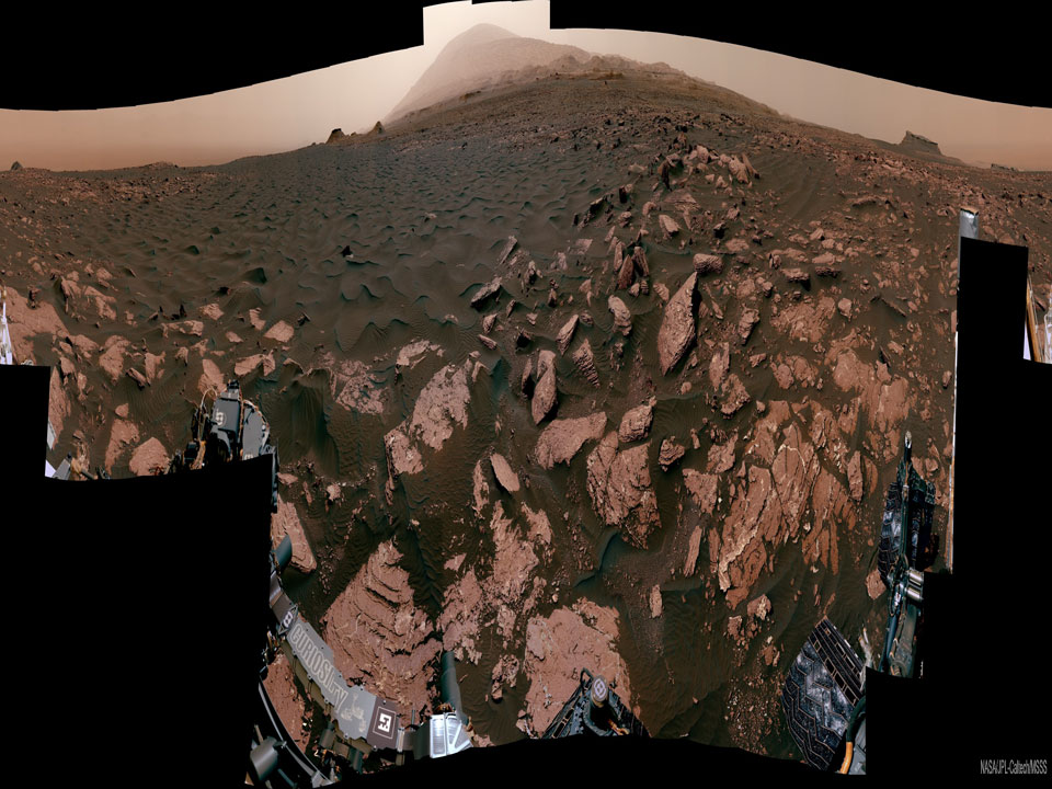 Das Bild ist horizontal verkürzt. Es zeigt hinten den Mount Sharp. Vorne ist Ogunquit Beach. Helles Gestein ragt aus dunklem Sand. Der Ort war auf dem urzeitlichen Mars manchmal von Wasser bedeckt und manchmal lag er an einem Strand.