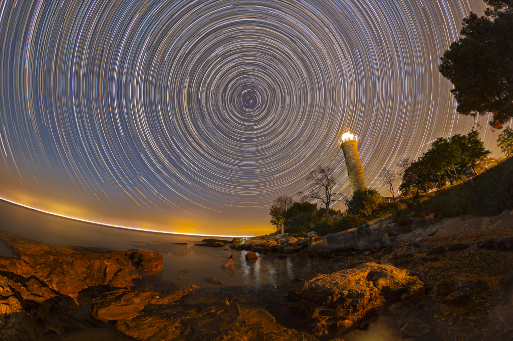 An der Küste der Halbinsel steht ein Leuchtturm, der Seglern beim Navigieren hilft. Am Himmel steht der Polarstern, um den scheinbar alle Sterne kreisen. Vorne ist eine felsige Küste.