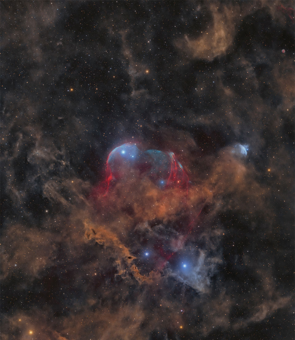 In einem Sternenfeld mit ein paar schwachen hellbraunen Staubwolken liegt in der Mitte eine große Wolke mit braunem Staub und blauen Gashüllen.