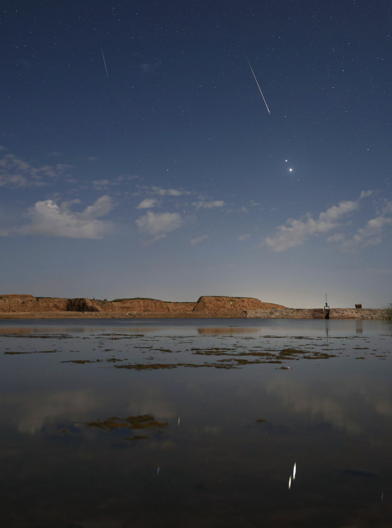 Am mondhellen Himmel stehen zwei gleißend helle Planeten eng beisammen. Sie spiegeln sich im Vordergrund in einem Gewässer. Ein Meteor der Perseïden fliegt scheinbar auf sie zu. In Wirklichkeit ist der Meteor viel näher.