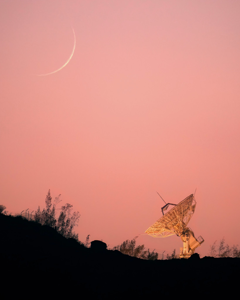 Am rosaroten Himmel über den Kanarischen Inseln leuchtet eine schmale Mondsichel knapp über dem Horizont. Unten ist eine Radioschüssel auf den Mond gerichtet.