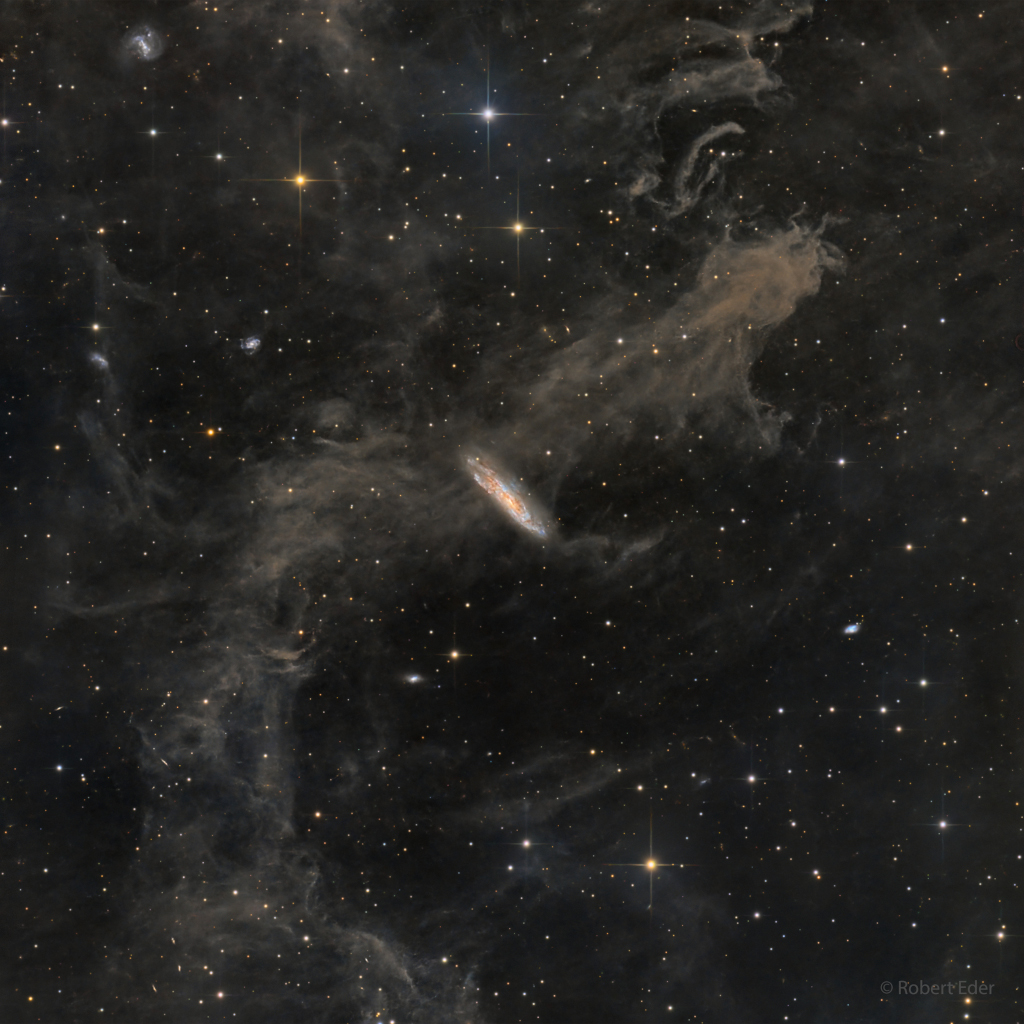 Vor einem dunklen Sternenhintergrund zieht eine graubraune Wolke von links unten nach rechts oben durch das Bild. Mittendrin eine kleine leuchtende Spiralgalaxie. Ein Klick auf das Bild lädt die höchstaufgelöste verfügbare Version.