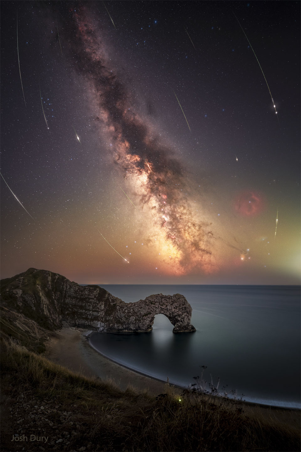 Über der Durdle Door an der englischen Küste bei Dorset steigt die Milchstraße steil über dem Meer auf. Vom Himmel fallen Meteore der Perseïden vom Himmel.