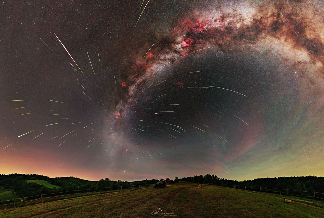 Über einer Wiese mit Hügeln am Horizont strahlt ein Sternhimmel. Das Band unserer Milchstraße zieht sich in einem Bogen nach rechts. Von einer Stelle der Milchstraße knapp über dem Horizont strömen viele Lichtspuren aus.