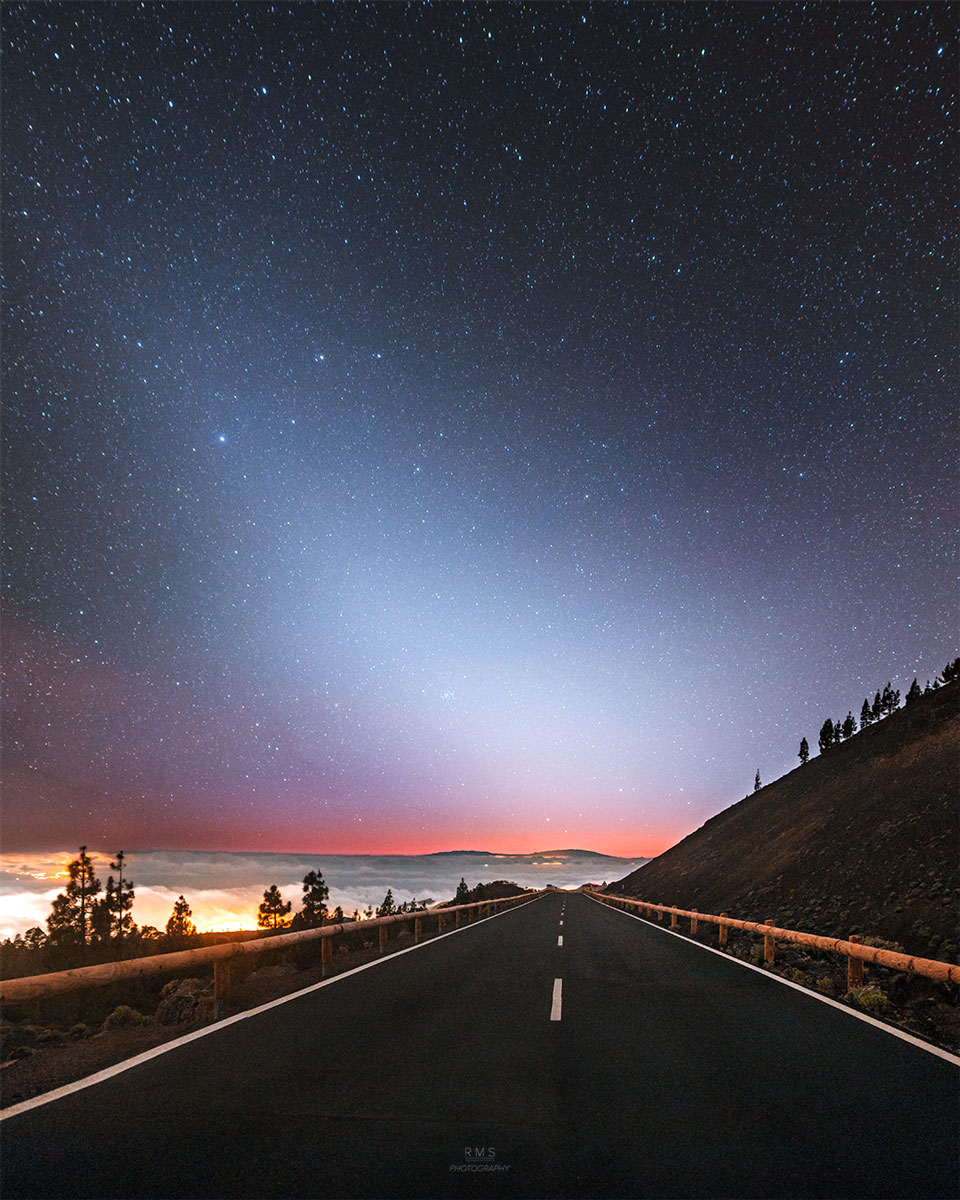 Die Straße im Bild führt scheinbar zu einem breiten Kegel aus Zodiakallicht. Der Sternhaufen M44 ist darin zu finden. Am Horizont ist noch Abendrot. Links sind Wolken, die von unten gelb beleuchtet sind.