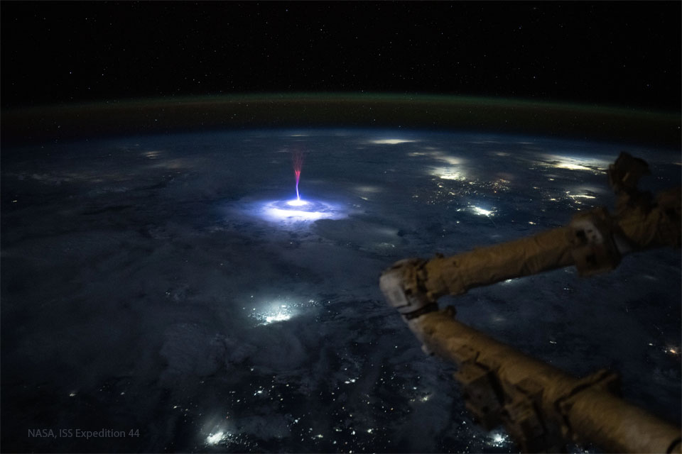 Blick auf die nächtliche Erde aus Sicht der Internationalen Raumstation. Im Vordergrund der Roboterarm der Raumstation, auf der Erde das Leuchten von Städten und ein nach oben weisender Blitz in Rot, Blau und Weiß.