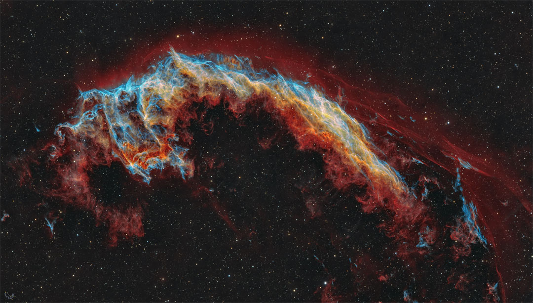 Ein leuchtender Nebel bildet einen zerfledderten Bogen. Er ist in roten und blauen Farben dargestellt und erinnert entfernt an eine Fledermaus. Der Nebel ist als NGC 6995 katalogisiert.