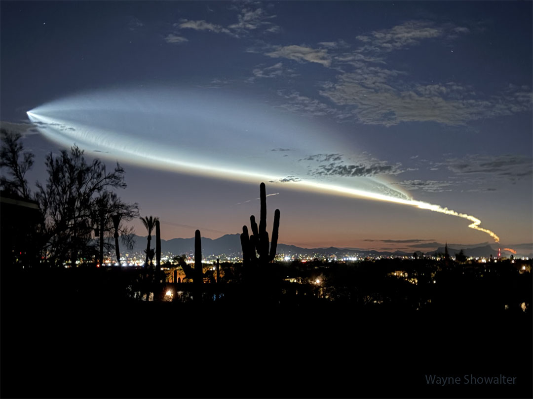 Über einer Landschaft mit den Silhouetten von Kakteen und den Stadtlichtern von Tucson startet eine Rakete in den Weltraum. Ihre Abgasfahne erinnert an einen riesigen Fisch. Sie wurde von der Sonne beleuchtet, die am Boden schon unter dem Horizont stand.