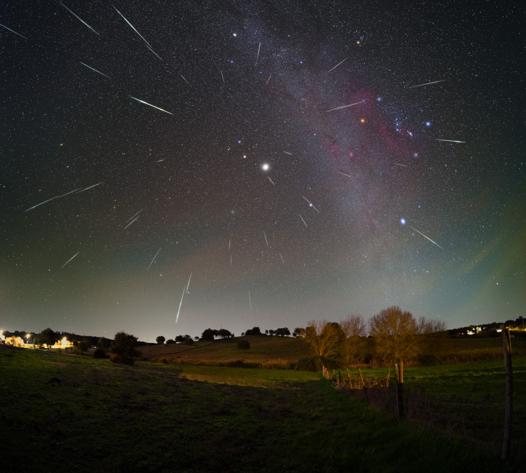 Über einer nächtlichen Landschaft in Portugal blitzen Meteore über den Himmel. Scheinbar strömen sie alle von einem Punkt in den Zwillingen aus, der nahe beim hellen Stern Kastor liegt. Bei Kastor und Pollux strahlt ein helles Licht, es ist der Planet Jupiter. Am Himmel sieht man außerdem die Milchstraße, rötliche Nebelwolken und ein Nachthimmellicht.