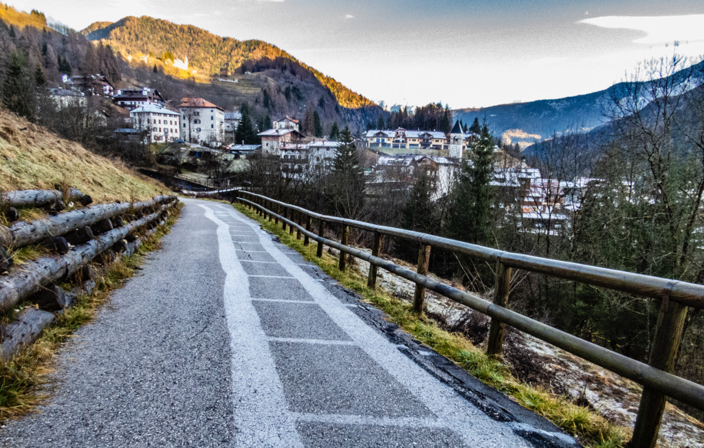 Vorne führt ein geteerter Weg zu einem Bergdorf, rechts ist ein Geländer. Auf dem Boden ist ein weißer Abdruck des Zauns. Dort, wo der Zaun einen Schatten auf den Boden warf, konnte der Frost am Boden nicht schmelzen und bildete ein weißes Muster.