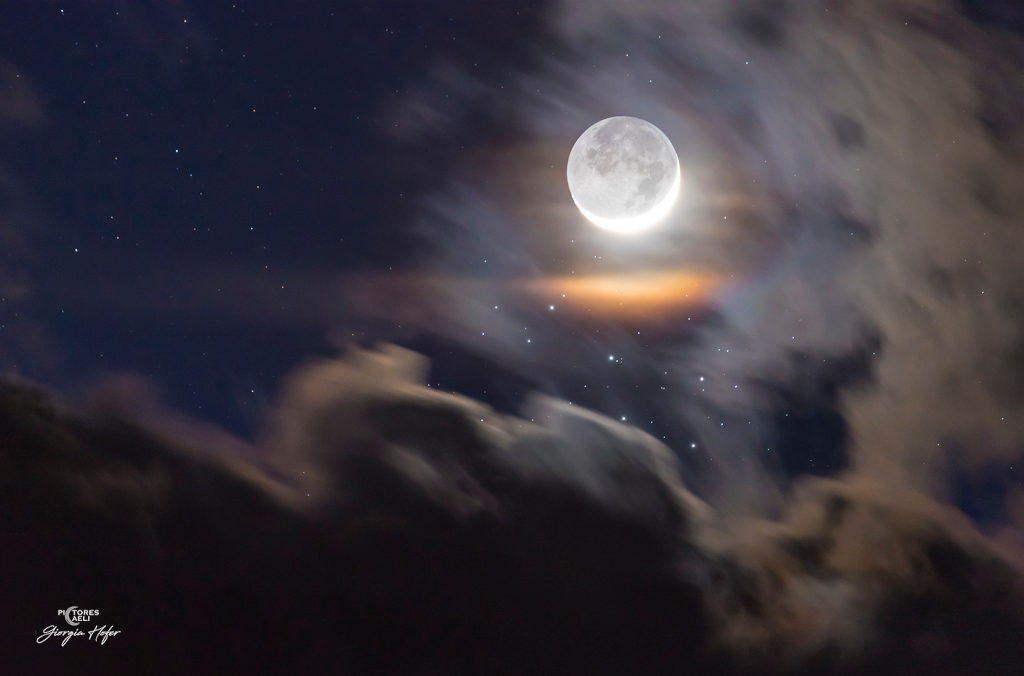 Der Sichelmond wird von der Erde beschienen. Wolken dämpfen das helle Licht. Dadurch sieht man die Sterne der Plejaden besser unter dem Erdtrabanten. Durch die Wolkenschleier entsteht eine leichte Mondkorona.