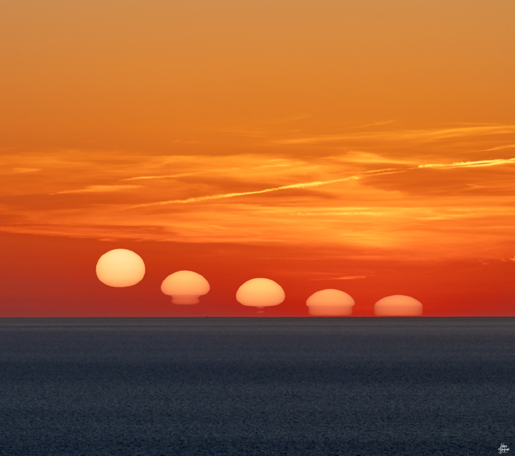 Die Sonne sinkt langsam zum Horizont. Der Himmel dahinter ist intensiv orange und rot gefärbt und von vielen Kondensstreifen überzogen. Die Sonne wirkt stark verzerrt und erinnert an Dalís schmelzende Uhren.