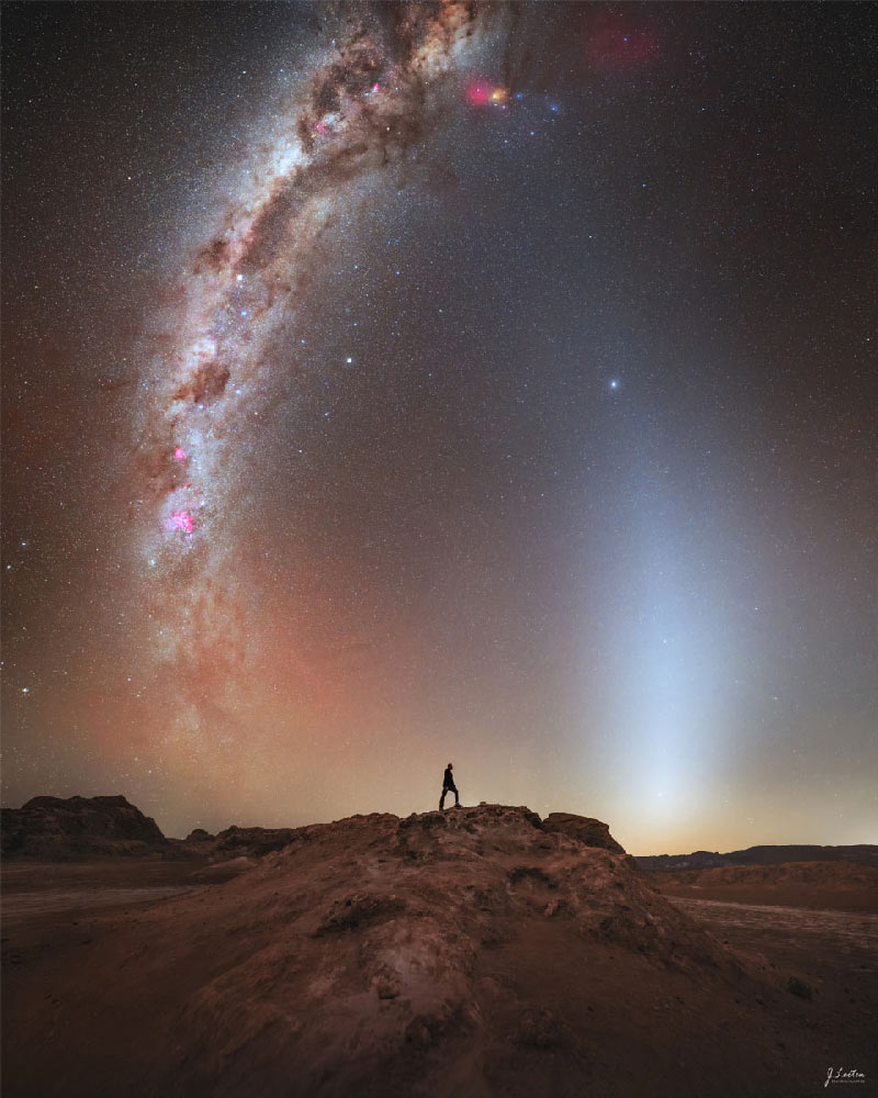 In einer felsigen Landschaft steht in der Ferne eine Person. Über ihr breitet sich der Sternenhimmel aus. Zu beiden Seiten der Person ragen helle, bogenförmige Lichtbänder vom Horizont auf.