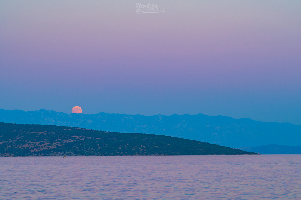 Über dem Meer ragen Berge der Insel Krk auf, über denen der Vollmond aufgeht. Zusammen mit dem Mond steigt auch ein bleigraues Band auf, das sich nach oben hin rosarot verfärbt. Es ist der Schatten der Erde, der oben in den Venusgürtel übergeht.