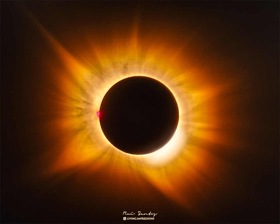 Die Sonne ist vom Neumond verdeckt. Daher strahlt ihre Korona in der Dunkelheit hell und farbenprächtig hinter dem Mond auf. Links ragt eine rote Protuberanz über den Mondrand.