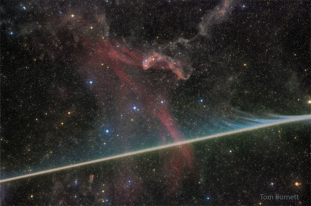 Eine helle Spur zischt quer durchs Bild. Dahinter leuchten blasse rote Nebelwolken am sternklaren Himmel im Sternbild Eidechse.