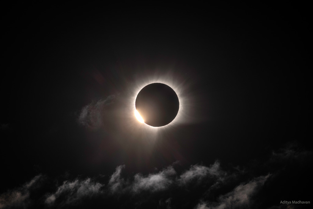 Der Mond verdeckt die Sonne, sodass der Himmel schwarz wirkt. Rund um den Mond schimmert die Korona. Links unten taucht die Sonne wieder auf, sodass der Diamantring leuchtet. Unten sind zarte Wolkenfetzen.