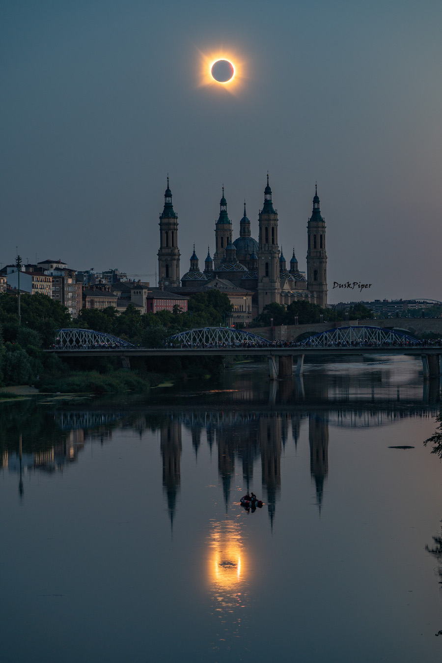 Über der Basílica del Pilar in Zaragoza (Spanien) leuchtet die Korona, weil die Sonne vom Mond verdeckt ist. Die Basilika hat mehrere pittoreske Türme. Finsternis und Basilika spiegeln sich vorne im Wasser des Ebro.
