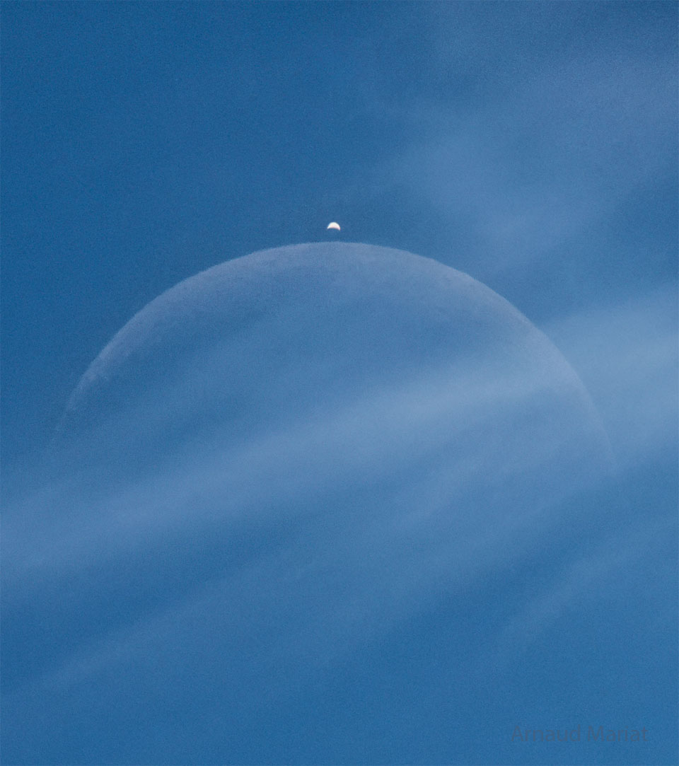 Hinter Wolkenfetzen schimmert die blasse Mondsichel. Darüber glänzt die helle Sichel der Venus. Die Sonne beleuchtet bei der Venus einen größeren Teil der Oberfläche als beim Mond. Der Himmel dahinter ist blau, denn die Bedeckung der Venus durch den Mond fand tagsüber statt.