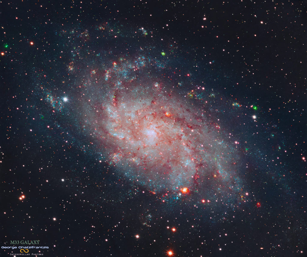 Die flauschige Spiralgalaxie M33 im Sternbild Dreieck liegt mitten im Bild. Rötliche Sternbildungsregionen und blaue Sternhaufen sprenkeln ihre losen Spiralarme. Um sie herum sind Sterne der Milchstraße verteilt. Am rechten unteren Rand leuchtet eine große rötliche Region.