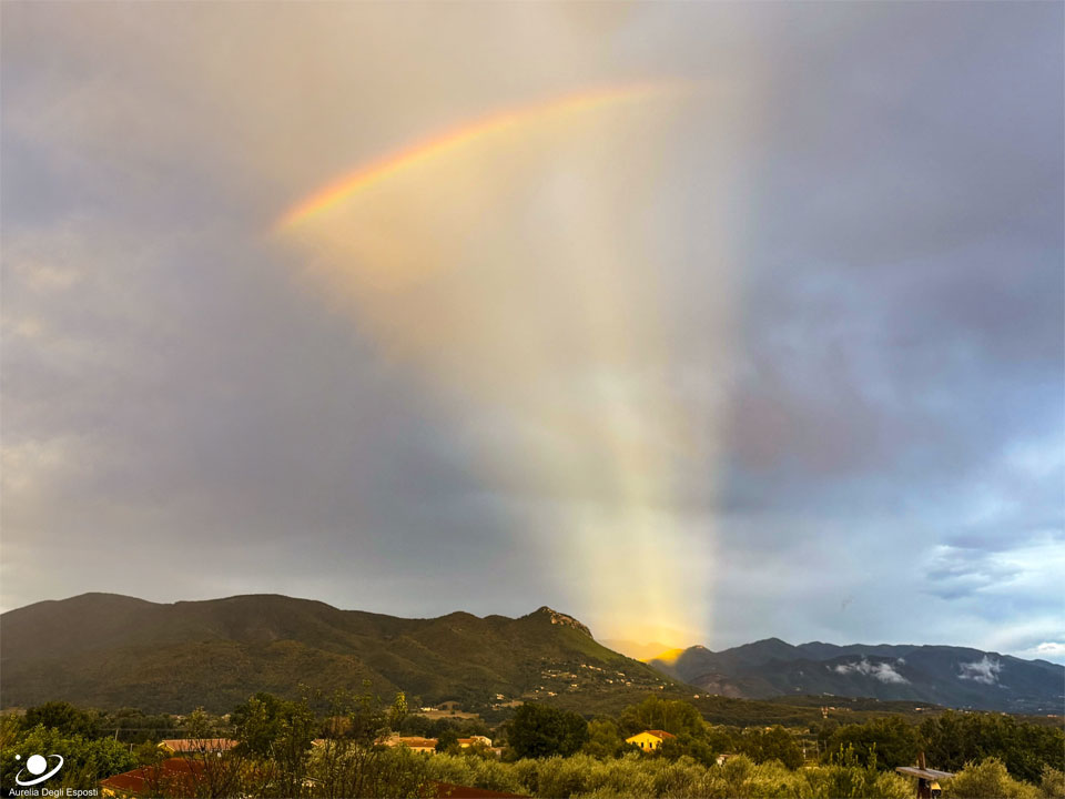Am Horizont ragen teils bewaldete Berge auf. Aus einem Einschnitt strahlt ein Fächer aus Licht nach oben, in dem ein Regenbogen schillert. Der Himmel ist von Wolken verhangen. Das Phänomen entsteht durch Gegendämmerungsstrahlen, kombiniert mit einem Regenbogen.