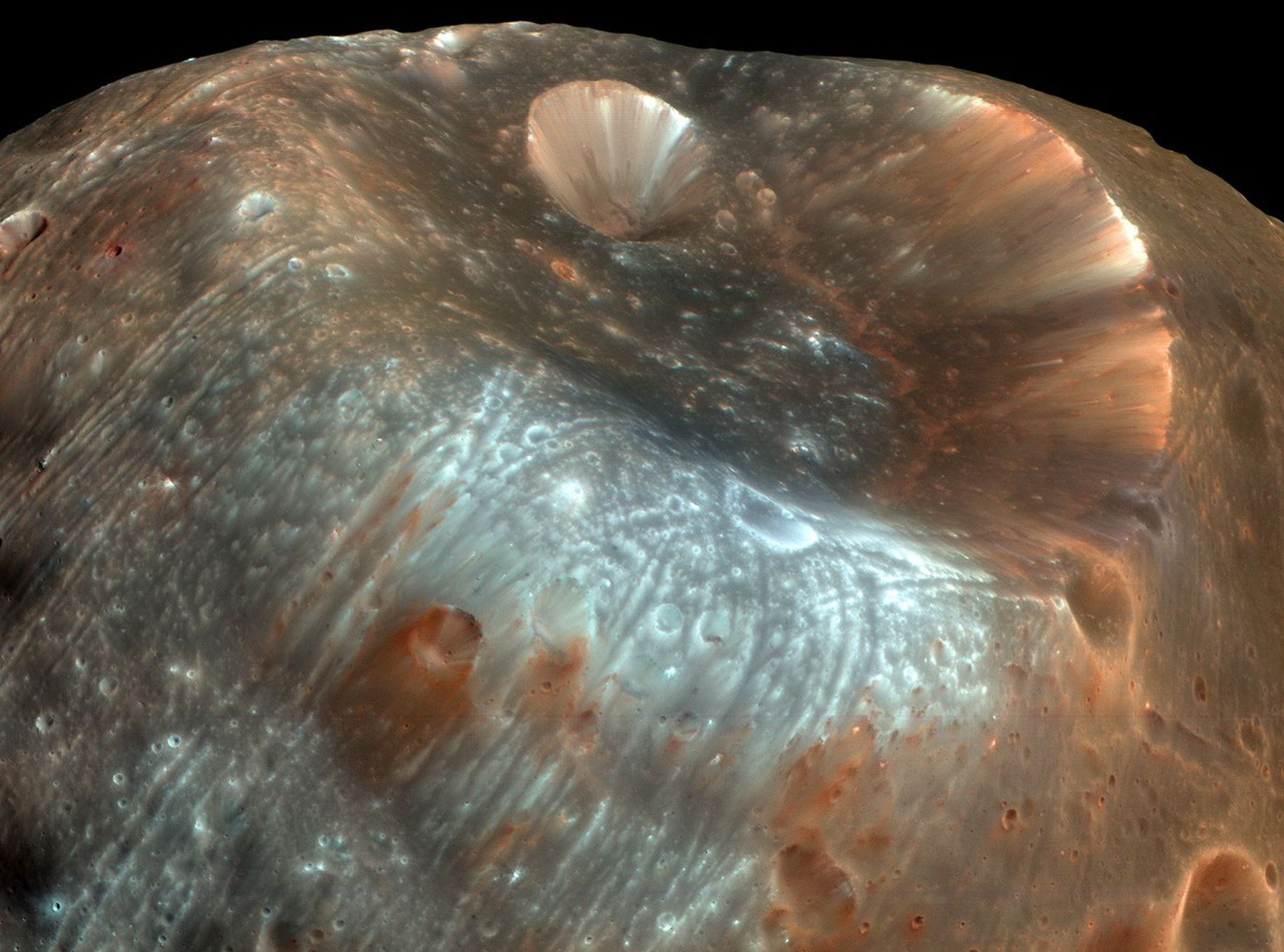 Ein asteroidenähnlicher Körper ragt ins Bild. Oben ist ein riesiger Krater, halb so breit wie der Mond Phobos. Links oben ist ein kleinerer Krater, der trichterförmig wirkt. Kleine Krater und viele Rillen überziehen den Mond. Rechts ist eine Kante, über die Geröll gerutscht ist. Dort befinden sich keine Krater. Unten ist helles Material, das von Kratern und Rillen überzogen ist. Vermutlich wurde dort kürzlich eine tiefer gelegene Schicht freigelegt.